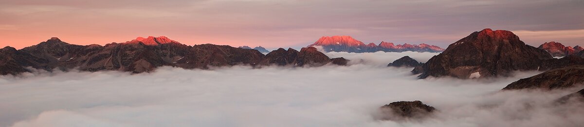 Vue panoramique des Alpes-Maritimes au coucher du soleil, avec les sommets baignés d’une lumière rosée et une mer de nuages recouvrant les vallées en contrebas ; à l’arrière-plan se distingue le massif de l’Argentera. Fin de la description de l’image.