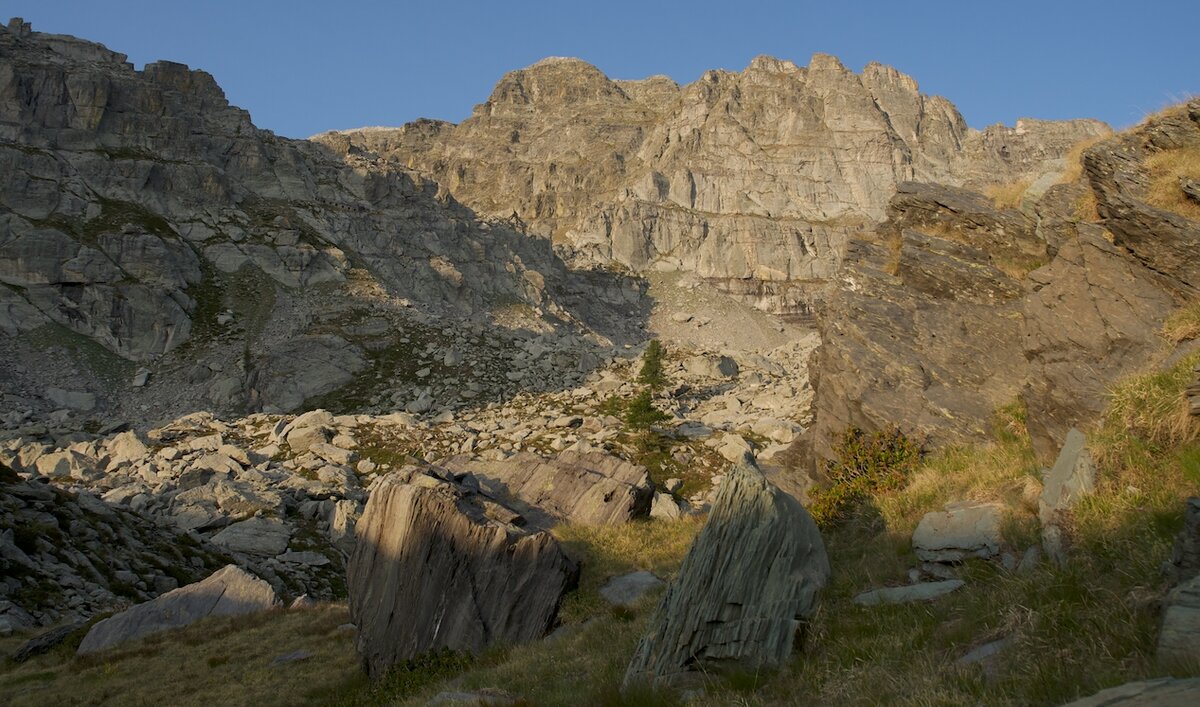 Vue du versant occidental du mont Bego dans la vallée de la Roya, avec des parois rocheuses éclairées par la lumière de l’après-midi et des blocs épars parmi les prés et les éboulis. Photo de G. Bernardi. Fin de la description de l’image.