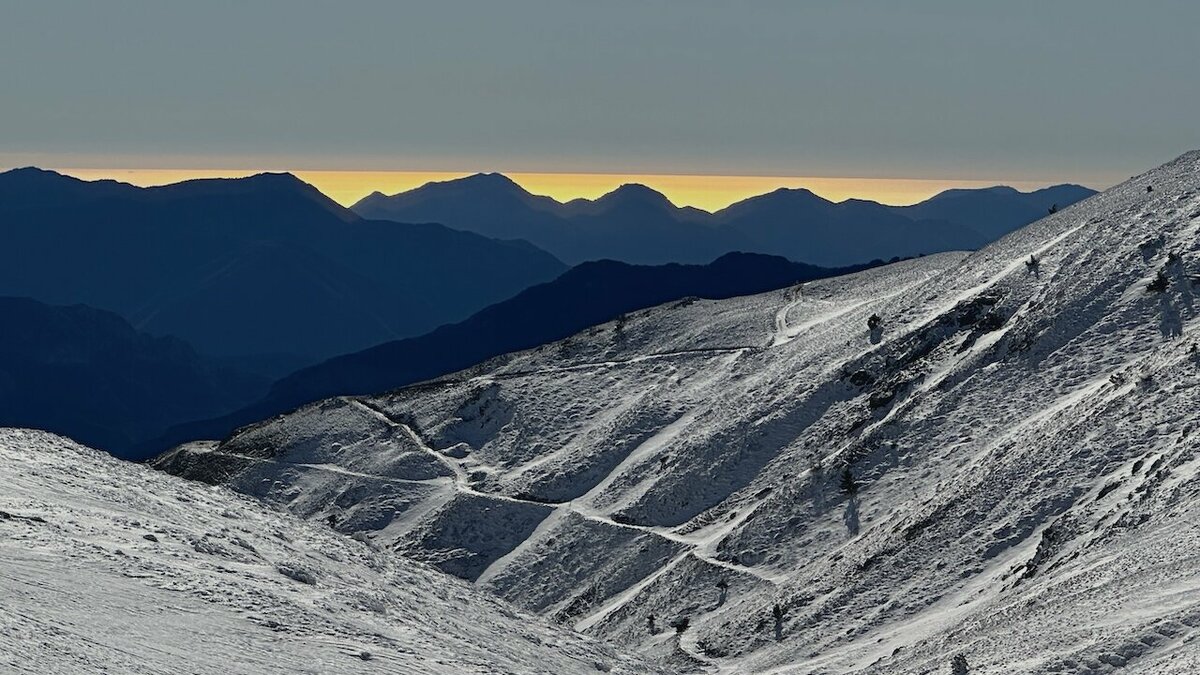 Vue hivernale des Alpes-Maritimes avec des pentes enneigées traversées par un sentier serpentant entre les rochers ; à l’arrière-plan, les silhouettes sombres des montagnes au coucher du soleil et une fine bande de lumière dorée à l’horizon. Archives APAM. Fin de la description de l’image.